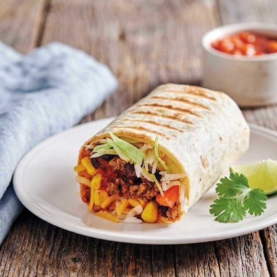 Grilled flour tortilla burrito filled with seasoned beef, rice, beans, cheese, corn, lettuce, and tomato, served with lime and cilantro.
