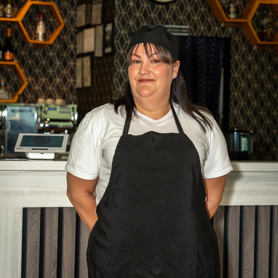 Restaurant staff member wearing a black apron and cap stands behind the counter, surrounded by patterned walls and honeycomb shelves.