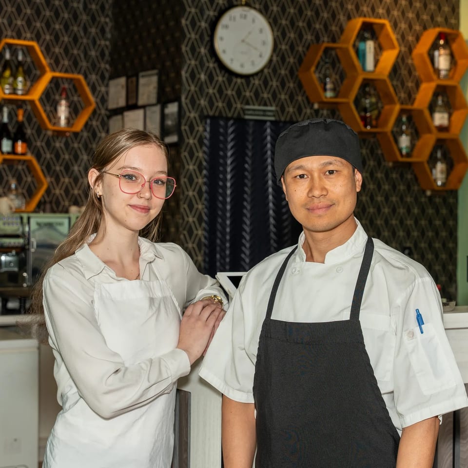 Restaurant chef and colleague pose behind the counter, with honeycomb shelves, bottles, patterned wallpaper, and a wall clock behind them.