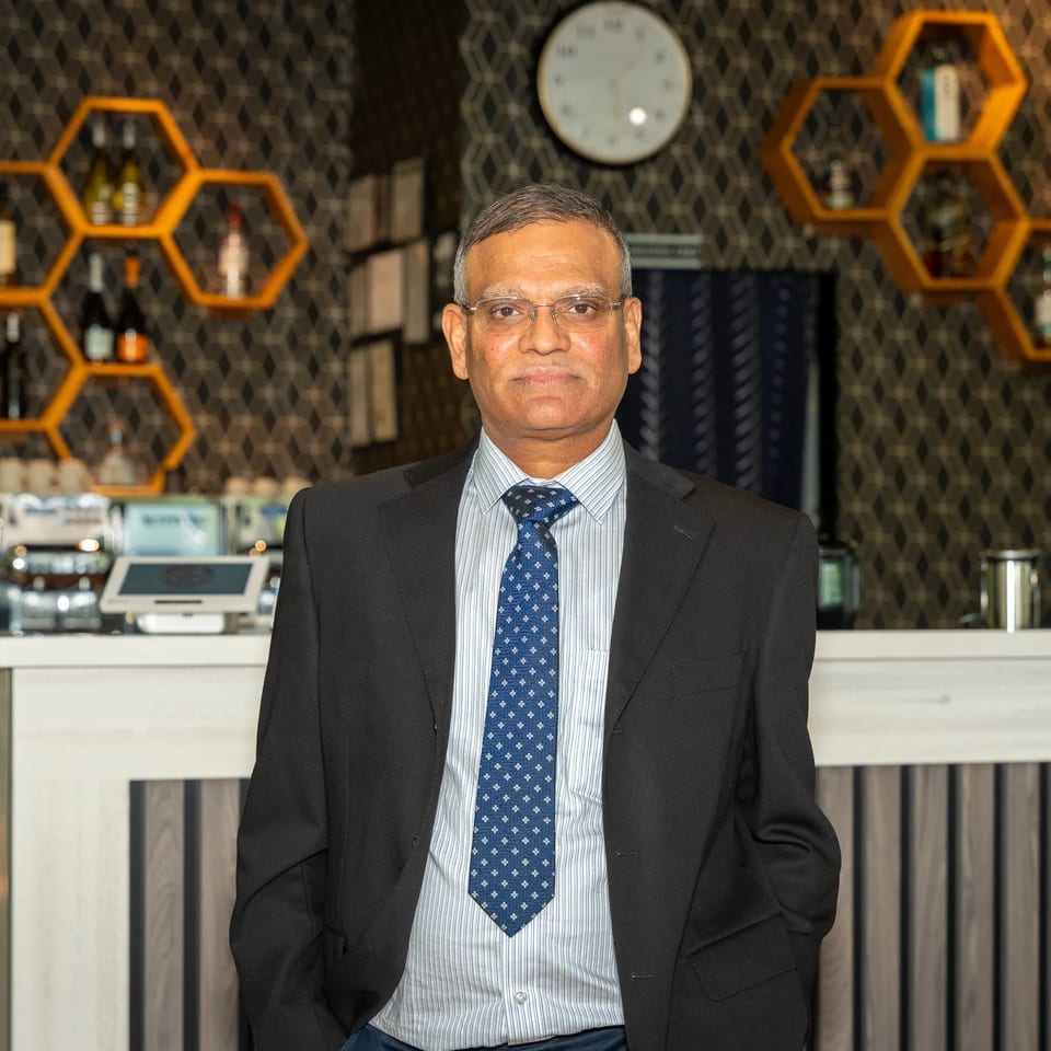 Man in a suit and tie standing behind a restaurant counter with honeycomb shelves and bottles.