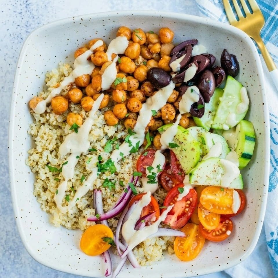 Quinoa bowl topped with roasted chickpeas, olives, zucchini, tomatoes, red onion, herbs, and creamy dressing.