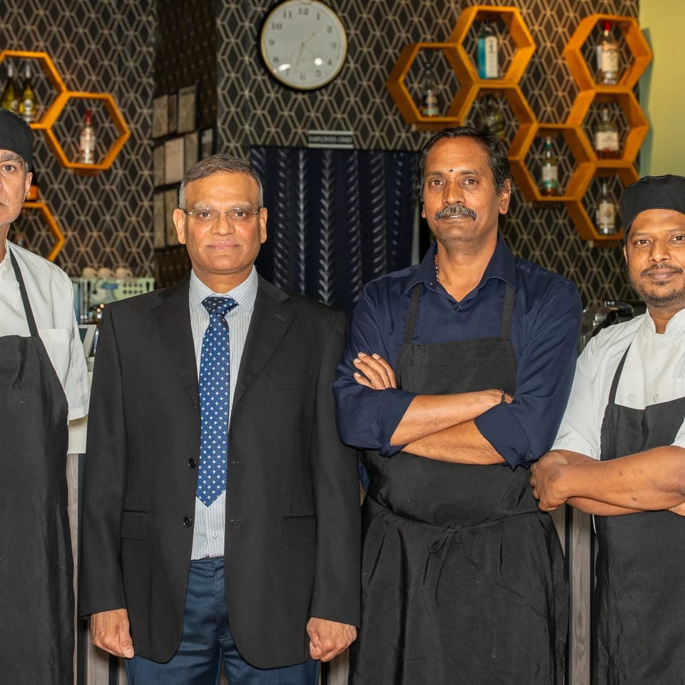 Four restaurant staff members pose together, including two chefs in white uniforms and aprons, against honeycomb shelves and patterned wallpaper.