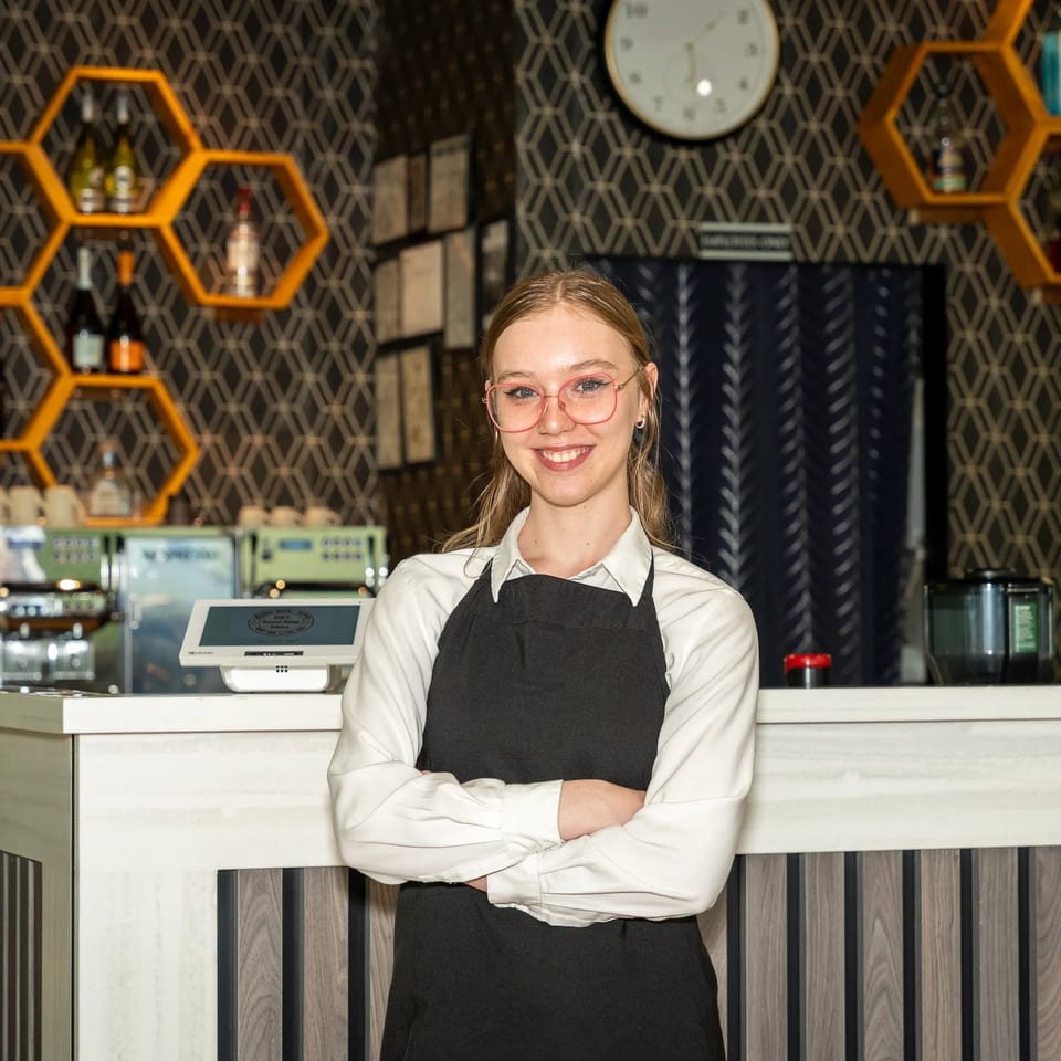 Smiling café employee in an apron stands behind the counter, surrounded by honeycomb shelves, bottles, and coffee equipment.